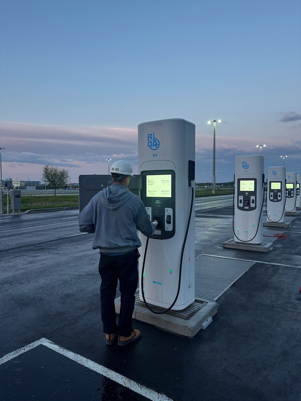 Electrician commissioning a row of DC fast chargers at a new charging plaza at dusk